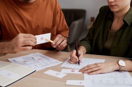 Male tutor of Chinese language pointing at paper card with hieroglyph while sitting in front of student recognizing and ticking it on test paper