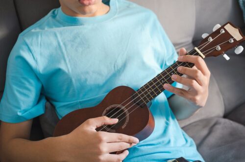A person in a light blue t-shirt plays a wooden ukulele while sitting on a couch, practicing for ukulele lessons in Singapore for beginners.