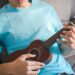 A person in a light blue t-shirt plays a wooden ukulele while sitting on a couch, practicing for ukulele lessons in Singapore for beginners.