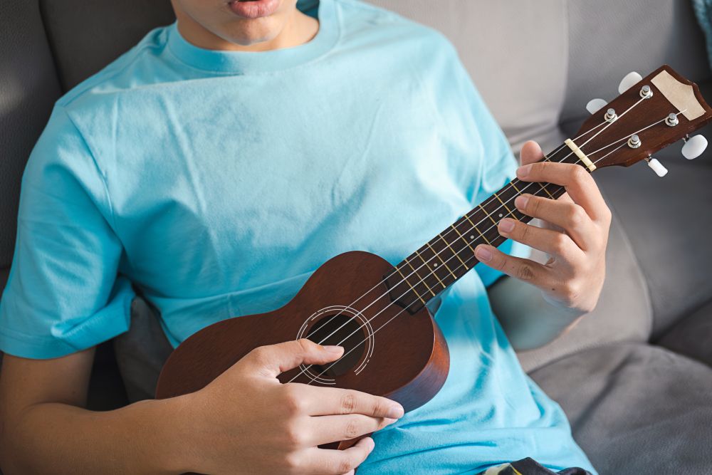 A person in a light blue t-shirt plays a wooden ukulele while sitting on a couch, practicing for ukulele lessons in Singapore for beginners.